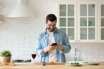 Handsome millennial single man cooking in modern kitchen holds smart phone searching recipes on...