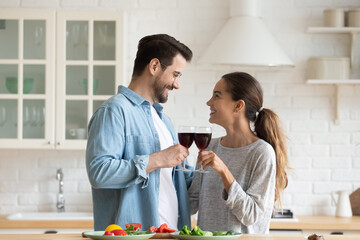 Happy young couple standing in cozy kitchen holding raising glasses of red wine celebrate house-warming or life events, enjoy romantic date at modern house, clinking wineglasses spend weekend at home