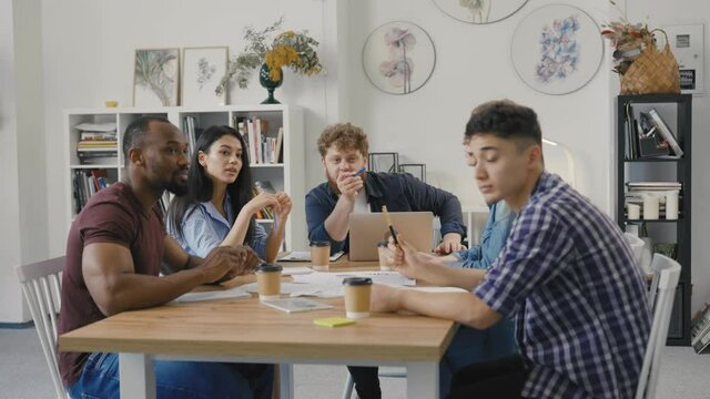 Friendly creative diverse colleagues team discuss startup project. Multiracial coworkers group sitting at table in modern office and discussing business issues in negotiation meeting. Slow motion.