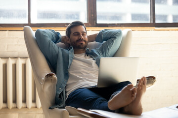 Serene millennial 30s man accomplish work on laptop put hands behind head relaxing on cozy leather...