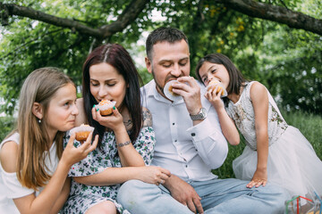Smiling parents with two cute daughters in summer clothes eating homemade cupcakes during picnic time at green garden. Concept of people, leisure and relaxation.