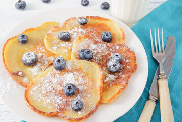 pancakes with blueberries on a plate and powdered sugar , glass of milk on a light  table,  blue napkin, fork,  knife,
