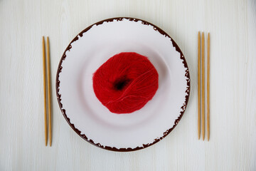Funny ball of yarn on white plate with wooden double pointed needles. Knitting as hobby. Ball of red mohair woolen thread on porcelain plate, top view, horizontal. Contemporary art concept