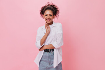 Curly girl in white shirt is smiling on pink background. Cheerful woman in denim skirt happily...