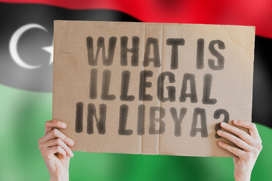 The question " What is illegal in Libya? " on a banner in men's hand with blurred Libyan flag on the background. Not allowed. Prohibitions. Outlaw. Rules. Policy. Law
