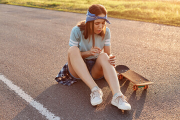 Young adult dark haired woman sitting on asphalt road after falling down from skateboard, injured her knee, feeling pain, looking at her leg with frowning face. - obrazy, fototapety, plakaty