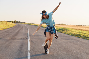 Fototapeta premium Full length portrait of slim sporty woman doing tricks on a skateboard, spending active time alone, outdoor on street, raised arms, looking down with excited facial expression.