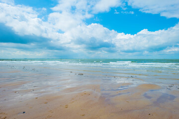 Sunlit waves on the yellow sand of a sunny beach along the North Sea illuminated by the light of a colorful sun and a blue cloudy sky in summer, Walcheren, Zeeland, the Netherlands, July, 2021
