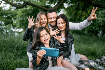 Fototapeta premium Cheerful parents and their cute daughters sitting together on green grass and using modern cell phone for taking self portrait. Concept of people, leisure and technology.