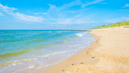 Obraz premium Sunlit waves on the yellow sand of a sunny beach along the North Sea illuminated by the light of a colorful sun and a blue cloudy sky in summer, Walcheren, Zeeland, the Netherlands, July, 2021 