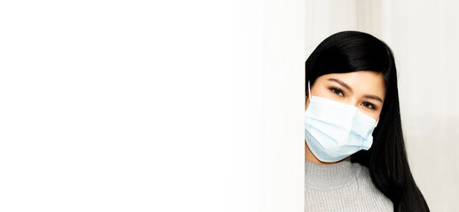 Asian woman with black hair wore mask and grey sweater stares at the camera from the corner in the white room.