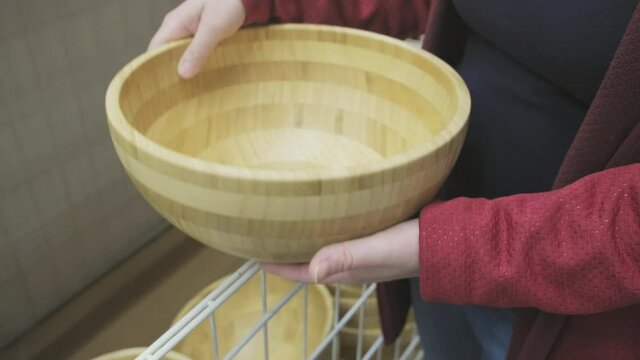 Caucasian woman in a store buys a large, round, deep, wooden plate made of natural wood. The concept of buying new dishes and kitchen equipment. Hands close up shot