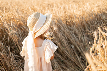 rear view of child girl in straw hat and beige muslin dress in wheat field on sunset