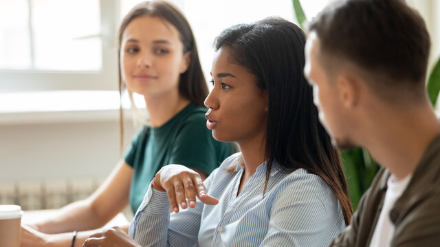 Serious mixed race employee, business team member speaking at corporate meeting, sharing ideas and solutions for project, talking to coworkers. Diverse group of interns discussing training task