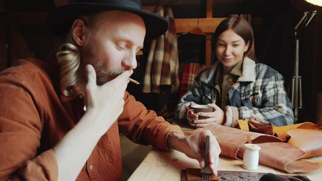 Young Caucasian craftsman smoking cigarette, speaking with wife and punching holes in leather with hammer and chisel at table in workshop