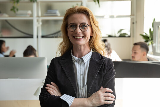 Happy senior business woman, leader, manager in glasses looking at camera with hands folded, ream working behind. Female mentor, coach, teacher posing in office with group. Head shot portrait
