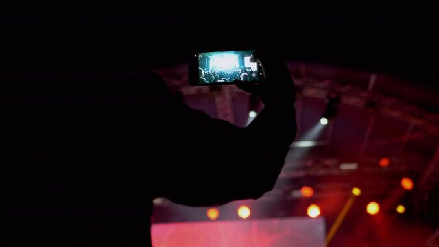 Male Record Video on a Smartphone of a Night Rock Concert on Open Stage. Fan is shoot a video, photo on a mobile phone. Stage lighting with spotlights. Flickering light. Crowd of people on screen. 4K.
