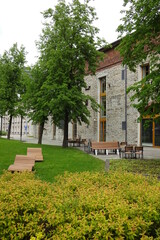 Curved wooden benches on green grass with bushes and trees around. Grey office building facade. Relax zone, place in the park. Ulemiste, Tallinn, Estonia