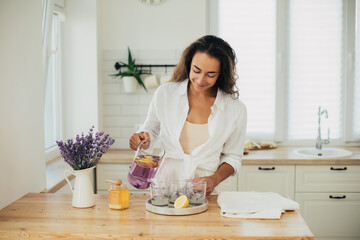 Young woman making lemonade in a kitchen of cozy house. Homemade healthy drink. - obrazy, fototapety, plakaty