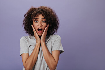 Curly woman in surprise looks at camera on purple background. Shocked brunette girl in grey tee posing on isolated backdrop