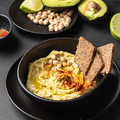 Black ceramic bowl with chickpea hummus on black background with chickpea beans , avocado, smoked paprika and lime multigrain bread,