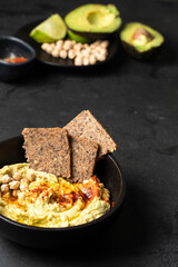 Black ceramic bowl with chickpea hummus on black background with chickpea beans , avocado, smoked paprika and multigrain bread