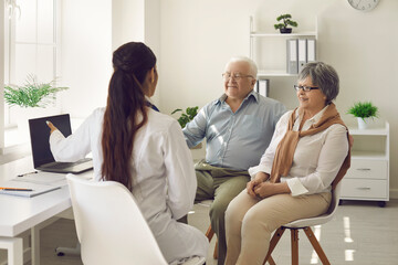 Happy mature couple and their doctor communicating while using laptop during medical appointment. Female doctor explains to senior patients test results pointing at laptop screen. Healthcare concept.