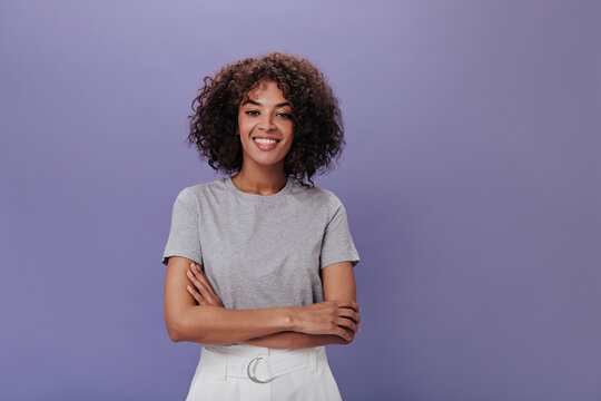 Portrait of young girl in gray t-shirt on purple background. Closeup snapshot of curly brunette woman in light outfit smiling on isolated backdrop