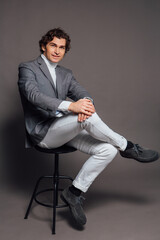 Tall handsome man dressed in white turtleneck, white jeans and grey jacket sitting on chair and posing on the grey background