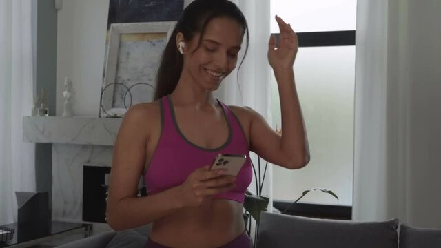 Young sports woman of a mixed race listens to music through wireless headphones. Submission with smartphone. Lady in a cozy living room at home. Woman dressed in sport pink top.