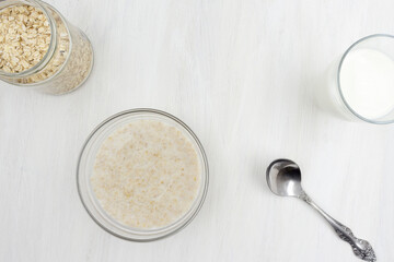 Oatmeal porridge cooked with milk, a glass of milk, a spoon, a plate on a white wooden table