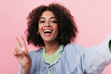 Joyful lady takes selfie and shows peace sign on pink background. Brunette woman in denim jacket smiling on isolated backdrop