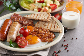 Traditional full English breakfast with fried eggs, sausage, tomato, beans, toast and bacon on a plate, Fresh fruit and vegetable salad, Milk and orange juice on stone background.