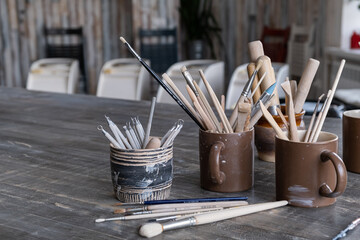 On the table in the creative workshop there are ceramic cups with brushes, metal and wooden stacks...