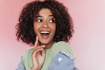 Joyful woman with curly hair laughs on pink background. Cheerful brunette girl in denim jacket smiling on isolated backdrop
