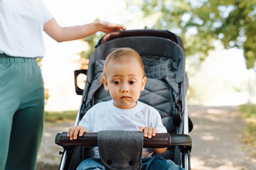 Portrait of a child with a funny emotion on face sitting in a stroller, outdoors. Mom's hand holding the handle of the stroller, walking with her little son on a summer day. Motherhood, family concept