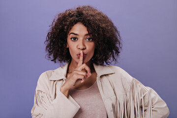 Brown-eyed woman in jacket with fringe asks to observe silence. Portrait of curly girl in beige...