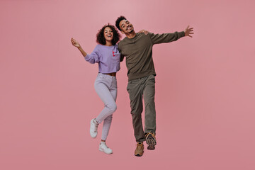 Happy couple in brown and purple outfits posing on pink background. Dark-skinned woman and brunette man smiling and hugging on isolated