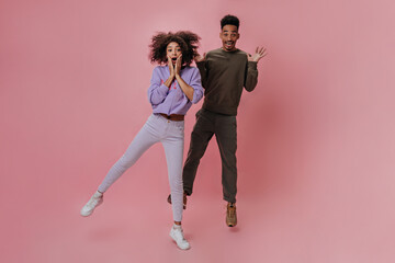 Surprised girl in purple sweater and jeans jumping with her boyfriend on pink background. Brunette woman looks shocked. Couple dancing on isolated