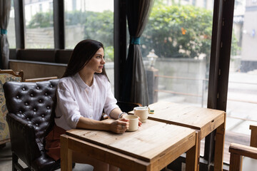 Young caucasian woman with long brunette hair wearing casual clothing and sitting in cafeteria holding coffee mug while looking away. Middle aged woman drinking tea while thinking about life or work