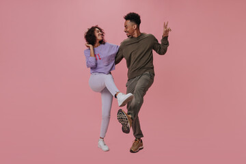 Brunette man shows peace sign and hugs his dark skinned girlfriend. Curly girl in purple sweater and her boyfriend dance on pink background