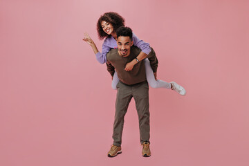 Couple in good humor having fun and showing peace sign on pink background. Brunette woman sitting on her boyfriend's back and smiling