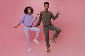 Positive couple in purple and brown outfits dancing on pink background. Dark-skinned man in sweater and curly woman in skinny pants move and have fun on isolated