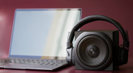 Obraz na płótnie Canvas A square black audio speaker and wireless headphones next to a slim modern laptop. Concept for digital music and wireless audio gadgets. Burgundy background. Daylight