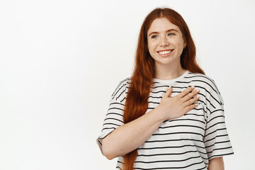 Image of happy girl with ginger hair, hold hand on heart and smiling, looking away with pleased and grateful face expression, thanking for smth, standing against white background