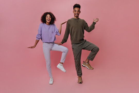 Couple having fun and dancing like Egyptians on pink background. Pretty woman in purple sweatshirt and man in brown pants standing in weird pose