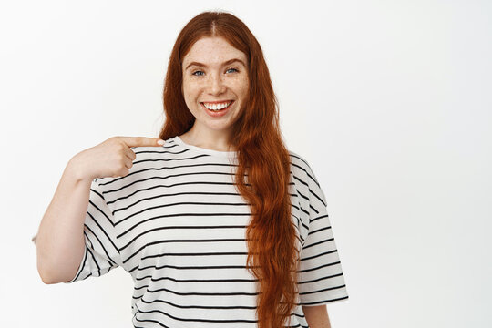 Image of smiling redhead girl with long natural hair, pointing at herself with pleased face expression, white teeth, showing something on her, standing against white background
