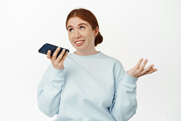 Image of redhead caucasian girl record voice message, talking on speakerphone, gesturing and chatting on mobile app, smiling relaxed, white background