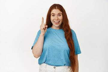 Eureka. Excited redhead girl with candid smile, pointing finger up, pitching an idea, got something interesting, showing advertisement on top, standing happy against white background
