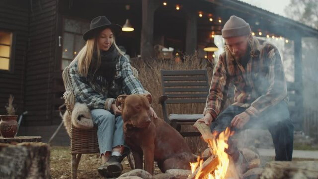 Man putting firewood in burning bonfire and sitting next to his wife as she petting pit bull dog in backyard of farmhouse